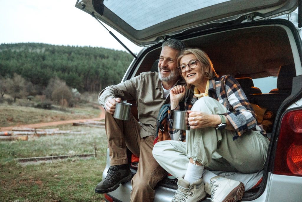 Older couple sitting in an open car trunk, smiling with mugs, forested hillside behind them.